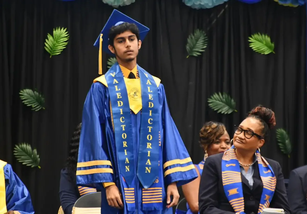Mirza N. Baig standing in cap and gown wearing two Valedictorian sashes as he is recognized during the Charlotte Amalie High School graduation ceremony, with faculty seated behind him.